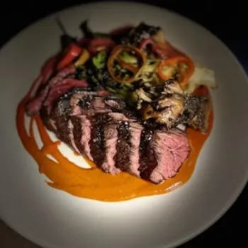 A plate of sliced medium-rare steak with grilled vegetables, served on a swirl of orange sauce.