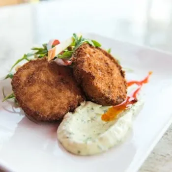 Two breaded and fried patties served on a white plate with a dollop of creamy sauce, garnished with greens and a streak of red sauce.