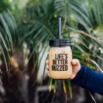 A hand holds a mason jar cup with a straw and the words "LIFE'S BETTER BUZZED" printed on it, in front of green palm leaves.