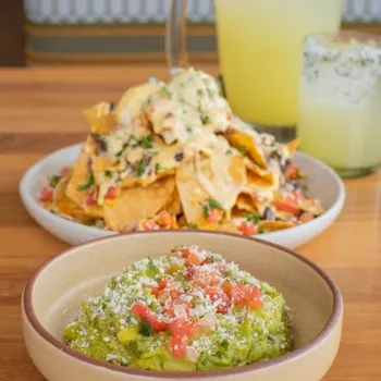 A bowl of guacamole with diced tomatoes and cheese in front of a plate of loaded nachos and a pitcher and glass of a yellow beverage on a wooden table.