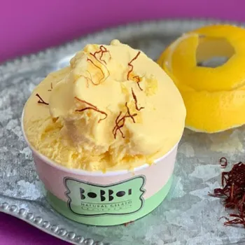 A cup of yellow gelato garnished with saffron threads sits on a silver tray next to a hollowed lemon and loose saffron strands.