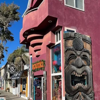 A red two-story building with a large carved wooden tiki mask at the entrance, located on a sunny street with nearby shops and palm trees.