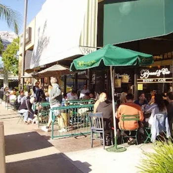 People are seated at outdoor tables under green umbrellas outside a café on a sunny day, with passersby walking along the sidewalk.