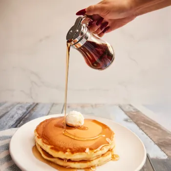 A hand pours syrup from a glass dispenser onto a stack of pancakes topped with a scoop of butter on a white plate.