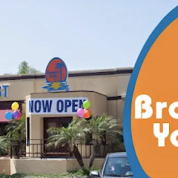 The exterior of Broken Yolk Cafe with a sign indicating it is now open, decorated with balloons and surrounded by palm trees.