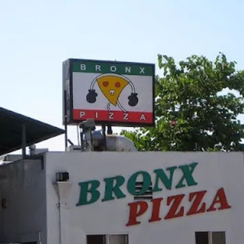 A sign on a building reads "Bronx Pizza" with a logo of a pizza slice flanked by two black circles, and trees are visible in the background.