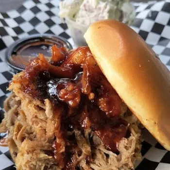 Pulled pork sandwich with barbecue sauce on a bun, served on checkered paper, with a side of coleslaw in a plastic cup.