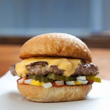 A cheeseburger with melted cheese, pickles, chopped onions, ketchup, and mustard on a bun, placed on a white surface.