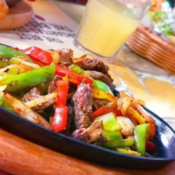 A sizzling plate of beef fajitas with sautéed green and red bell peppers and onions, served on a wooden board with drinks in the background.