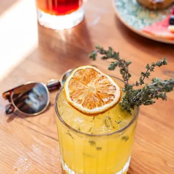 A yellow cocktail with ice, garnished with a dried citrus slice and herbs, sits on a wooden table beside sunglasses; another drink and food are blurred in the background.