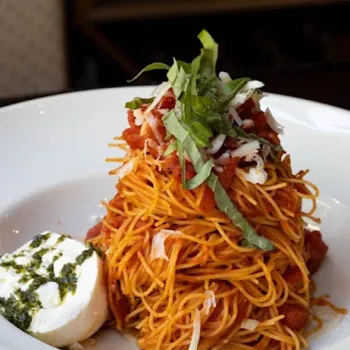 A serving of spaghetti with tomato sauce, topped with shredded basil and cheese, is plated next to a portion of mozzarella with pesto on a white dish.