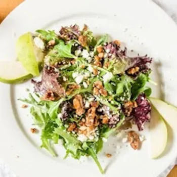 A white plate with mixed greens salad, crumbled cheese, candied nuts, and green apple slices sits on a white napkin next to a fork and salt and pepper shakers.