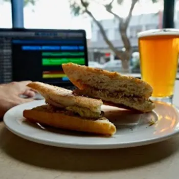 A plate with a sandwich is in front of a person using a laptop, with a glass of beer on the table and a street view visible through the window.