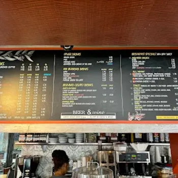 A large café menu board displays drink options, juice, breakfast specials, oatmeal bowls, and prices above a counter with staff and coffee machines visible below.
