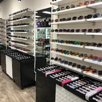 A retail store displays a wide variety of sunglasses on white wall shelves and black display tables, arranged in neat rows under bright lighting.