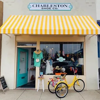 A storefront with a yellow and white striped awning, a teal door, display of clothes and hats in the window, and a yellow tricycle parked outside.