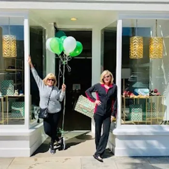 Two women stand in front of a store window display, one holding green and white balloons and a large bag, both smiling on a sunny sidewalk.
