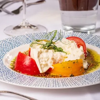 A plate of burrata cheese served with red and yellow tomatoes, olive oil, and herbs on a patterned dish, with a glass of water and wine in the background.