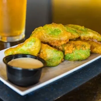 A rectangular plate with battered and fried avocado slices, a small cup of dipping sauce, and a glass of beer on a dark surface.