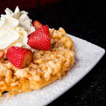 A funnel cake topped with sliced strawberries and a swirl of whipped cream, served on a white square plate with a dusting of powdered sugar.