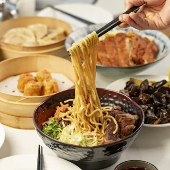 A hand uses chopsticks to lift noodles from a bowl of ramen, surrounded by dim sum, dumplings, eggs, and dipping sauce on a dining table.