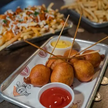 A tray with four mini corn dogs on skewers, served with ketchup and mustard; loaded fries and regular fries are blurred in the background.