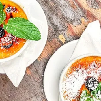 Two ramekins of crème brûlée on white plates, topped with mint leaves, berries, and powdered sugar, set on a rustic wooden table.