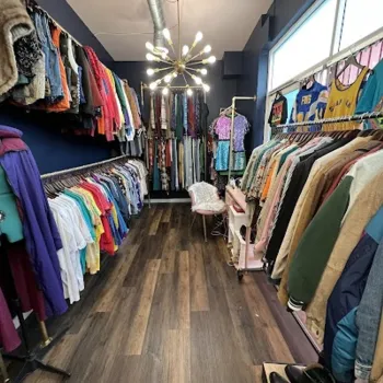 A narrow boutique with racks of colorful vintage clothing on both sides, a modern chandelier overhead, and a chair at the end of the aisle.