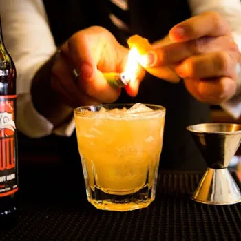A bartender uses a lighter to ignite an orange peel over a cocktail glass filled with an orange drink and ice, with a bottle and jigger nearby.