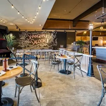 Restaurant interior with round wooden tables, metal chairs, a rickshaw, string lights, and a large "Shanghai Saloon" mural on the back wall.