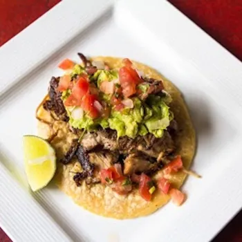 A taco topped with shredded meat, guacamole, diced tomatoes, and a lime wedge on the side, served on a white square plate.