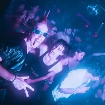 A crowd of people dance in a dimly lit nightclub with neon lighting; one person in front has spiked hair and sunglasses, making a hand gesture.