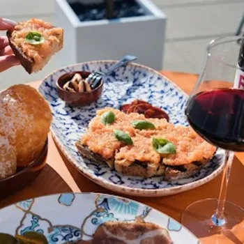 A table set with tomato-topped toast, fried bread, olives, cured meats, and a glass of red wine. A hand lifts a piece of toast. A bottle of wine is visible in the background.