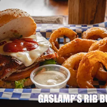 A rib burger with melted cheese, ketchup, lettuce, and tomato on a bun, served with onion rings and dipping sauce on a tray. Text reads "Gaslamp's Rib Burger.