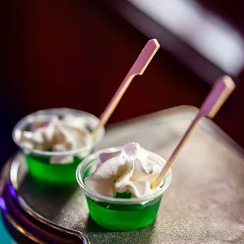Two small cups of green gelatin topped with whipped cream, each with a wooden stick, placed on a curved metallic surface.