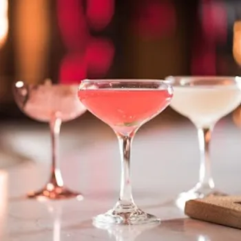 Three coupe glasses filled with different colored cocktails—pink, white, and pale—are arranged on a marble surface with a blurred background.