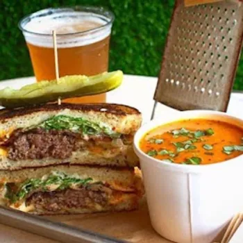 A grilled sandwich cut in half with a pickle on top, a cup of tomato soup, a pint of beer, a grater, and a salt shaker on a tray.