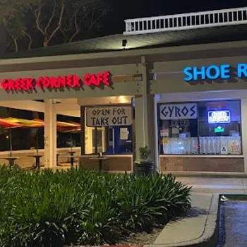 Nighttime view of a strip mall with signs for "Greek Corner Cafe" and "Shoe Repair;" the cafe displays "Open for Take Out" and "Gyros" signs in the window.