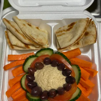 A takeout container with pita bread, sliced carrots, cucumbers, tomatoes, olives, and a serving of hummus in the center.