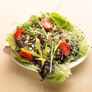 A white bowl filled with mixed greens, sliced cherry tomatoes, sprouts, seeds, and chopped vegetables sits on a light-colored surface.