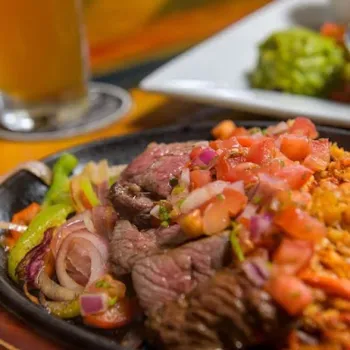 A plate of grilled steak topped with diced tomatoes and onions, served with sautéed bell peppers, onions, and a side of seasoned rice.