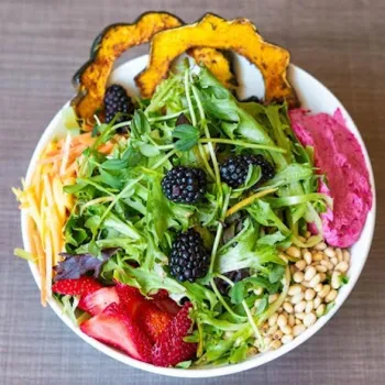 A bowl of mixed greens topped with blackberries, sliced strawberries, pine nuts, shredded carrots, roasted squash, and a scoop of pink beet hummus.