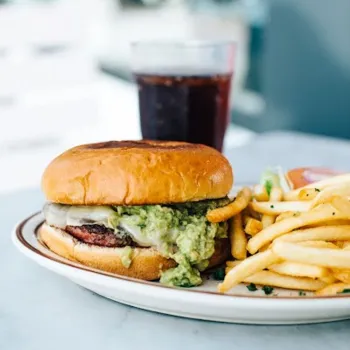 A plate with a burger topped with guacamole, a side of French fries, and a glass of dark soda on a light-colored table.