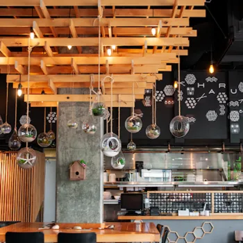 Modern restaurant interior with a wooden ceiling feature, hanging glass terrariums, a concrete pillar, open kitchen, and black wall with geometric white designs.