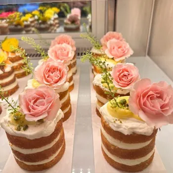 Mini layered cakes with white frosting, topped with pink artificial roses and green garnish, displayed on a refrigerated shelf.