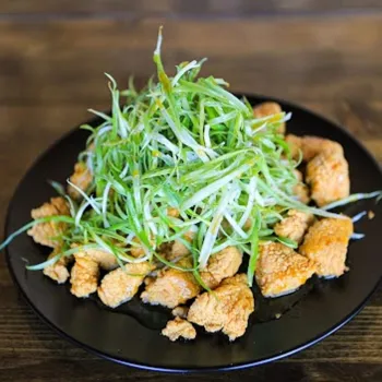 A black plate with pieces of fried chicken topped with a large pile of thinly sliced green onions, set on a wooden surface.