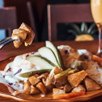 A fork holds a bite of roasted potatoes above a plate with more potatoes, pear slices, and a sauced dish, with an orange drink in the background.