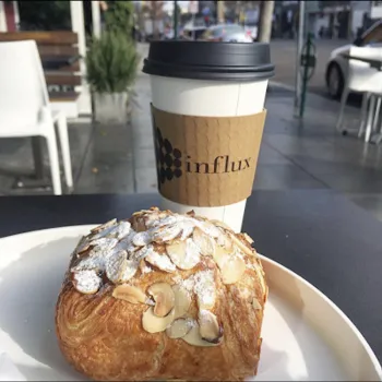 A pastry topped with sliced almonds and powdered sugar on a plate, with a to-go coffee cup labeled "influx" in the background on an outdoor table.