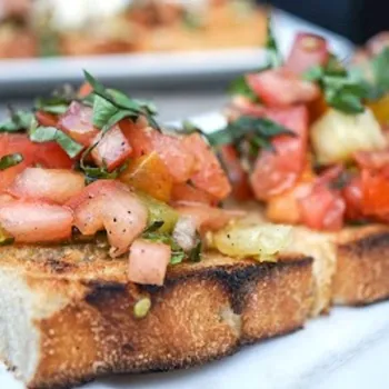Close-up of toasted bread topped with diced tomatoes, basil, and seasoning, served on a white plate.