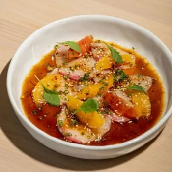 A white bowl containing sliced fish, orange segments, green herbs, and seeds in a brown sauce, served on a light-colored wooden surface.